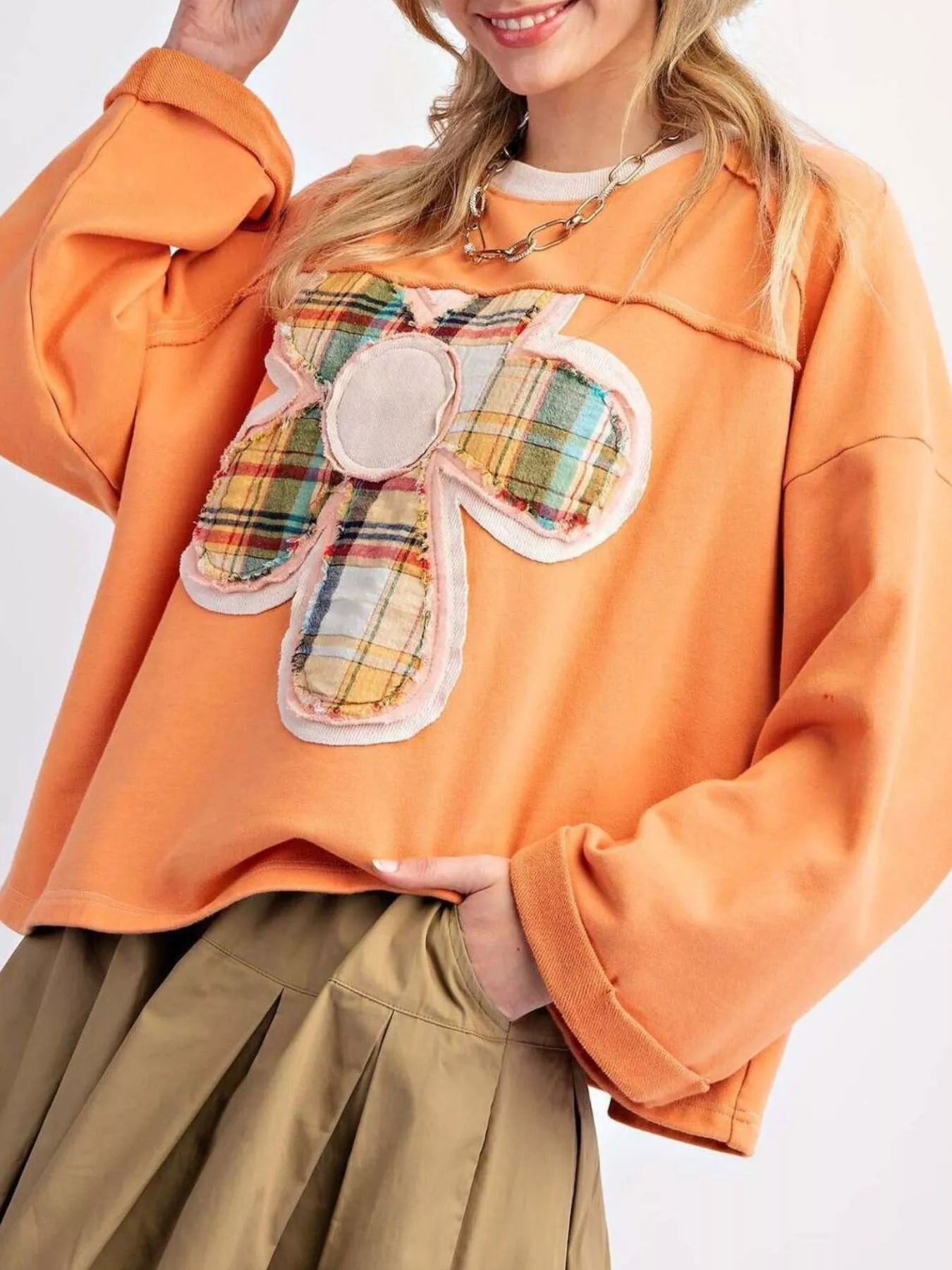 Bright orange pullover with plaid flower appliqué, styled with a pleated khaki skirt.
