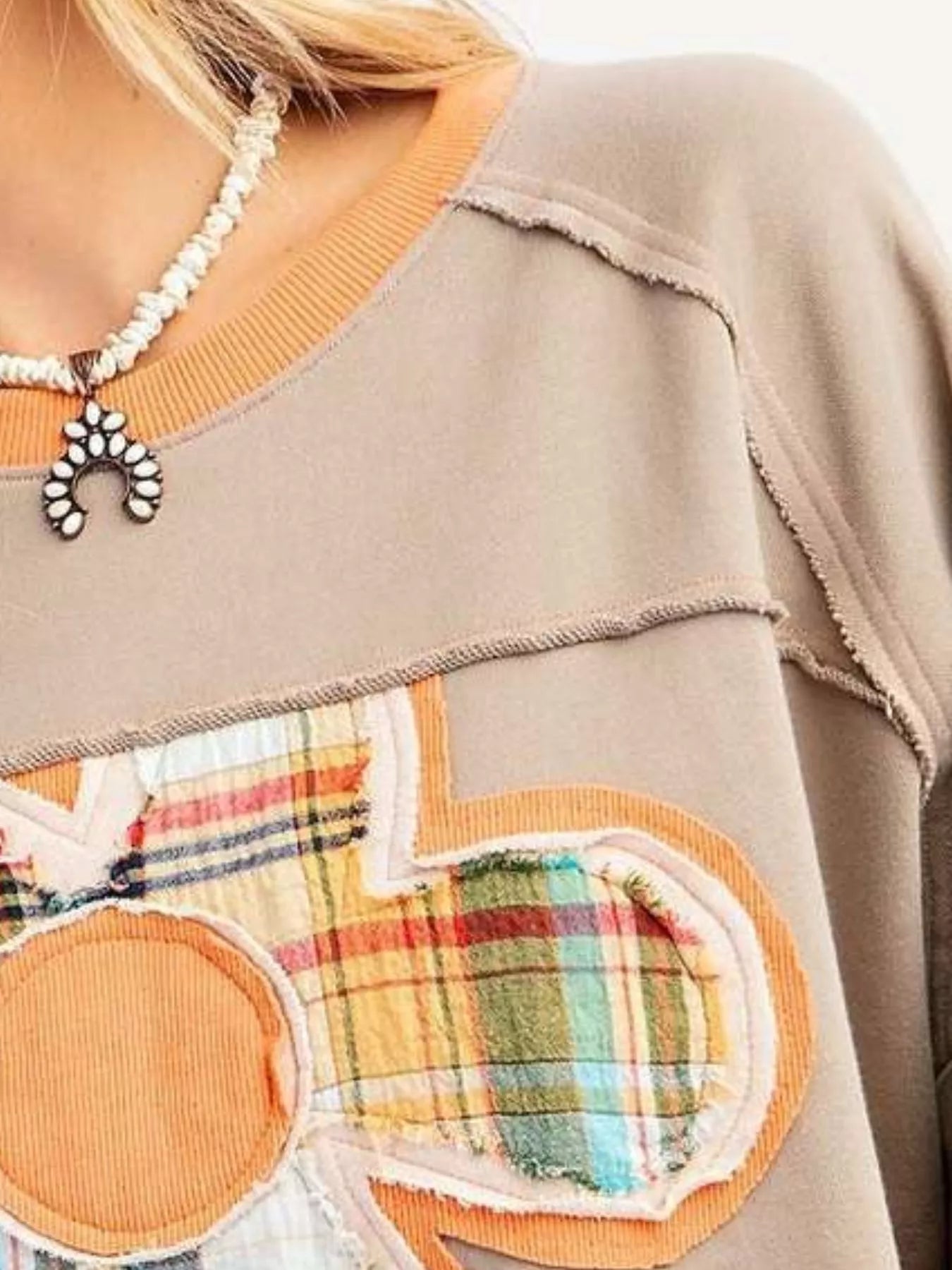 Taupe pullover with oversized plaid flower appliqué in orange tones, styled with light pink pants.