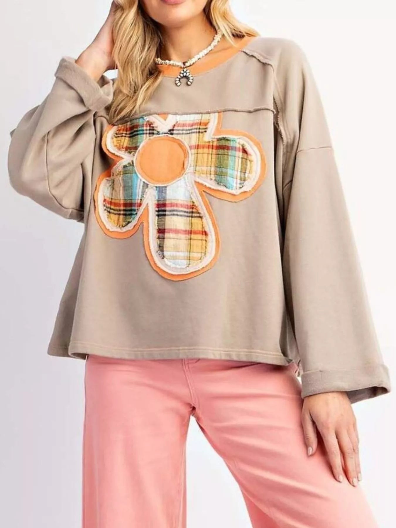 Taupe pullover with oversized plaid flower appliqué in orange tones, styled with light pink pants.