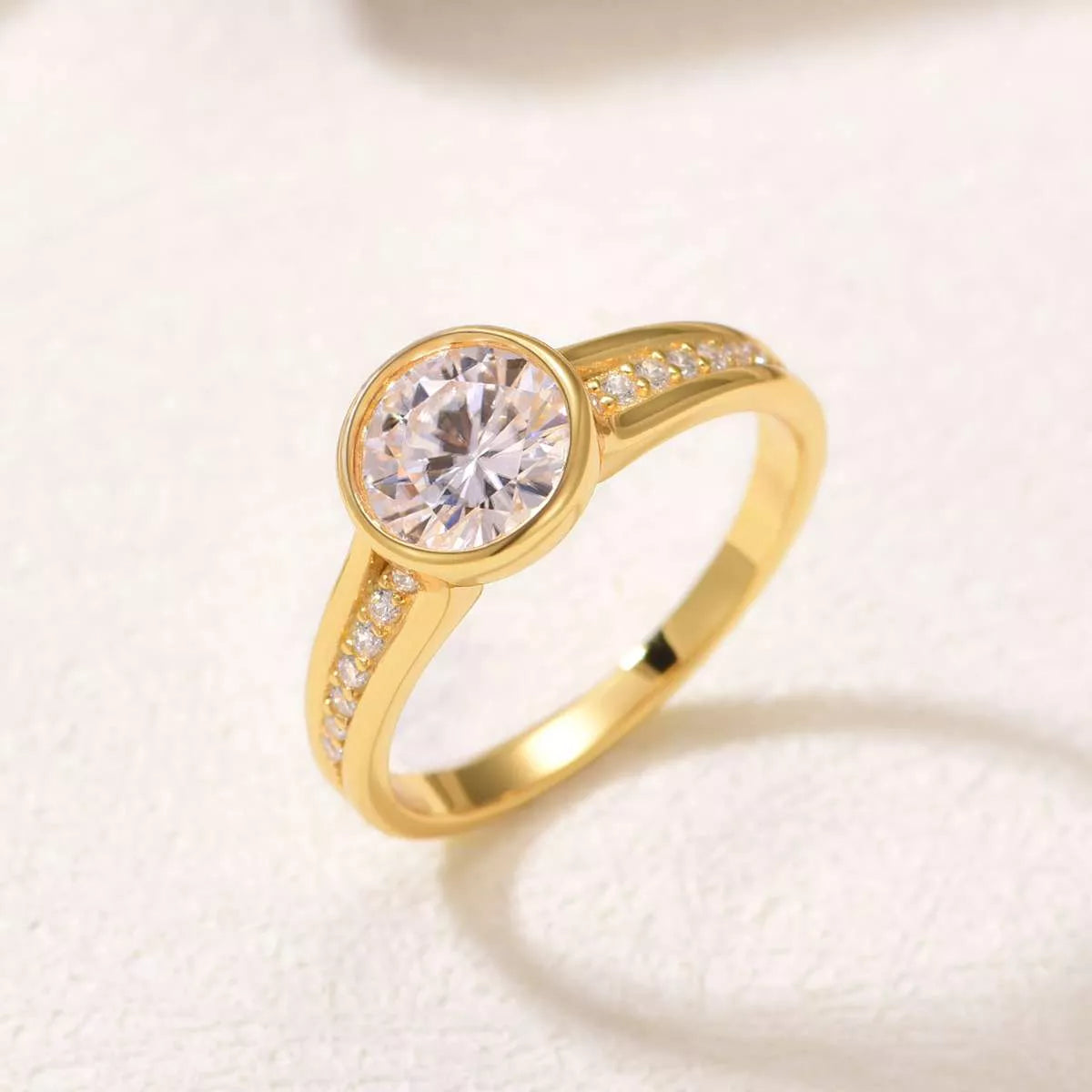 Close-up of a gold bezel-set moissanite ring with side zircon accents displayed in a blush jewelry box.