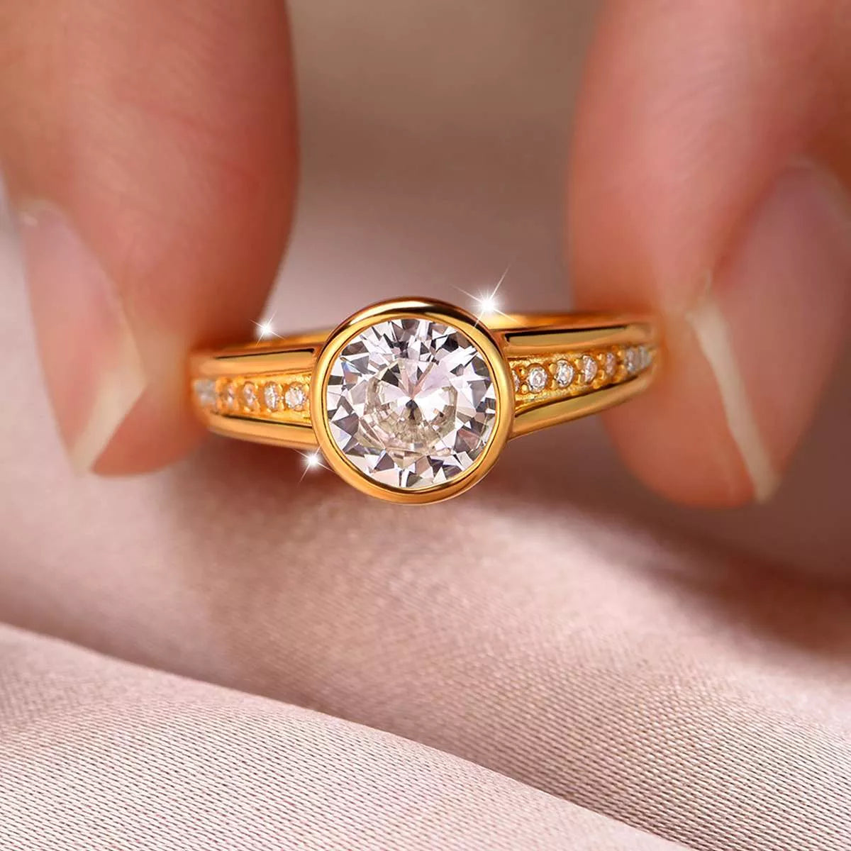 Close-up of a gold bezel-set moissanite ring with side zircon accents displayed in a blush jewelry box.
