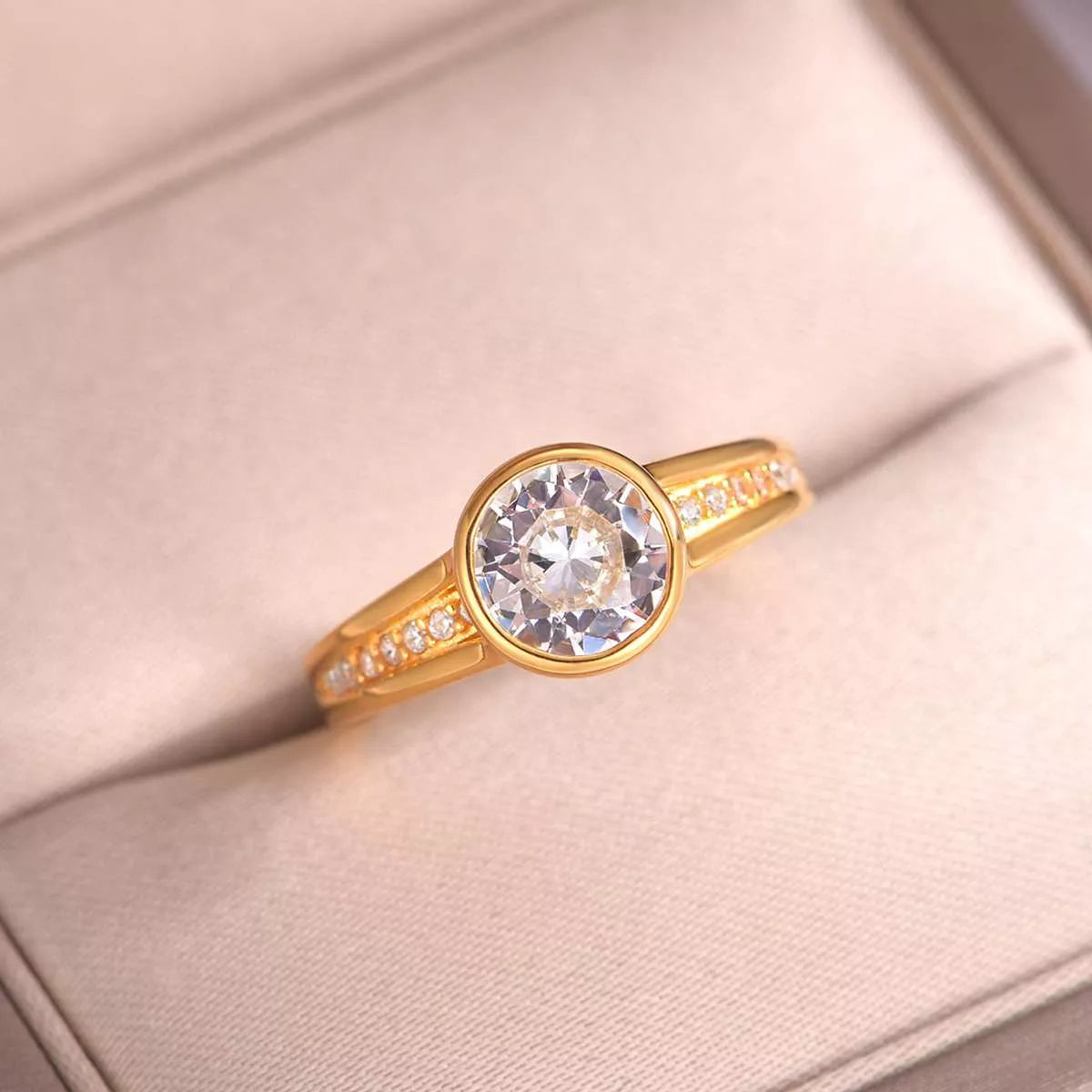 Close-up of a gold bezel-set moissanite ring with side zircon accents displayed in a blush jewelry box.
