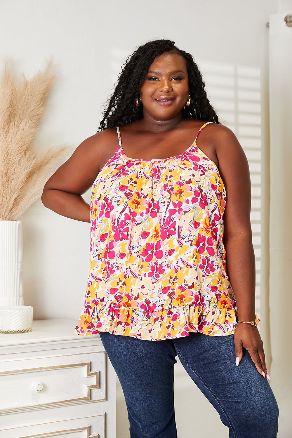 Model wearing a bright floral ruffle hem cami top with thin straps and a scoop neckline, paired with dark jeans.