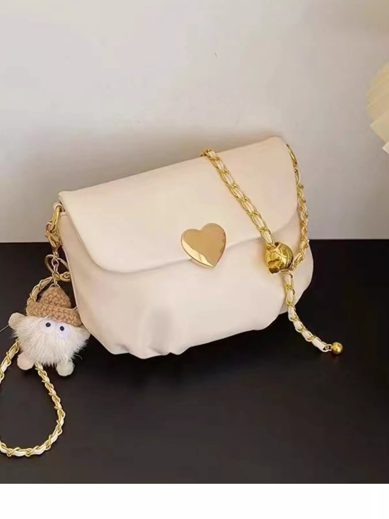 Cream mini crossbody bag with gold heart clasp and chain strap resting on beige fabric.
