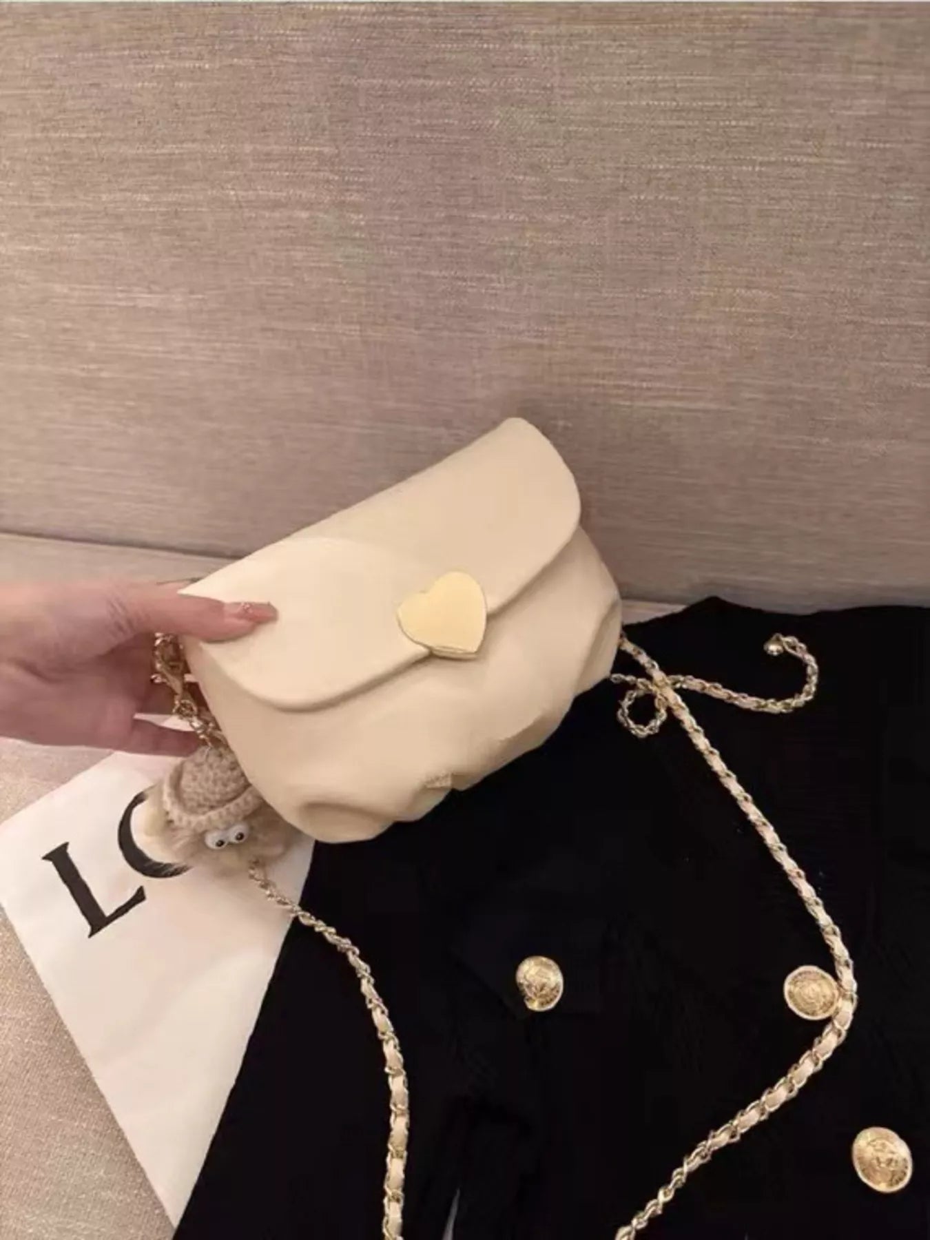 Cream mini crossbody bag with gold heart clasp and chain strap resting on beige fabric.