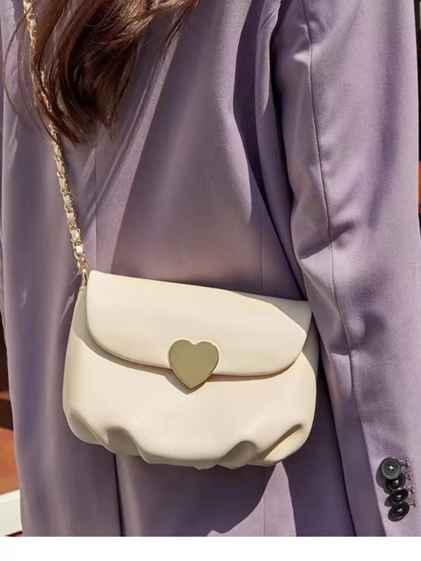 Cream mini crossbody bag with gold heart clasp and chain strap resting on beige fabric.