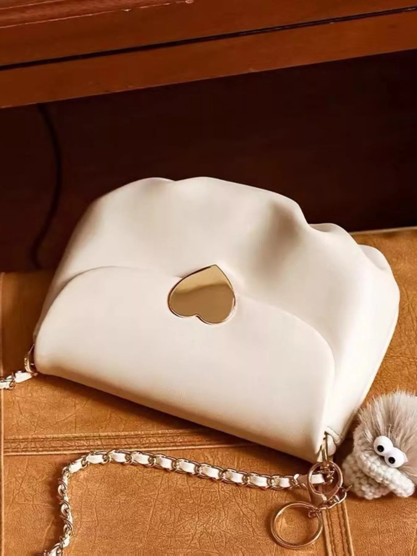 Cream mini crossbody bag with gold heart clasp and chain strap resting on beige fabric.