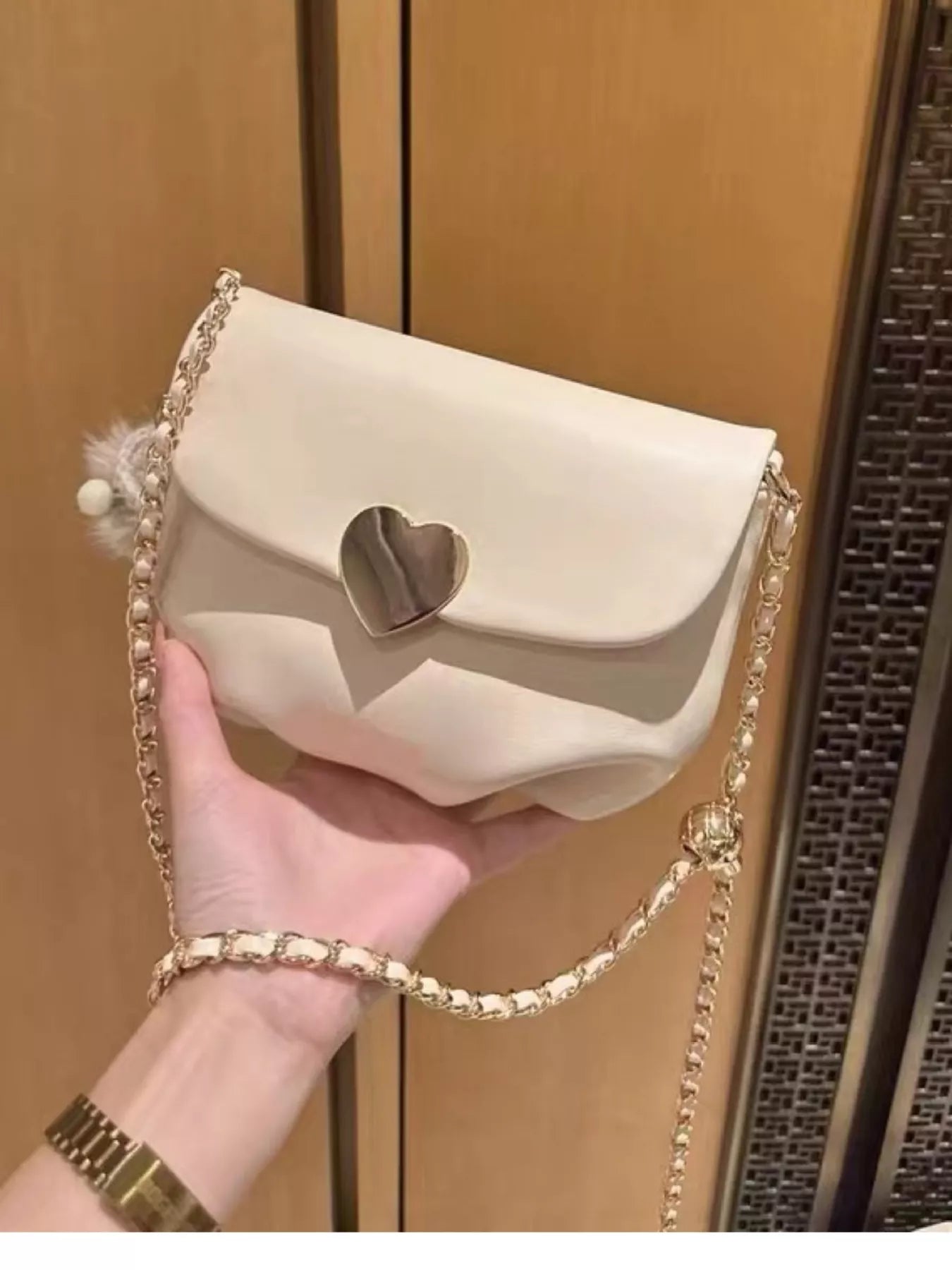 Cream mini crossbody bag with gold heart clasp and chain strap resting on beige fabric.