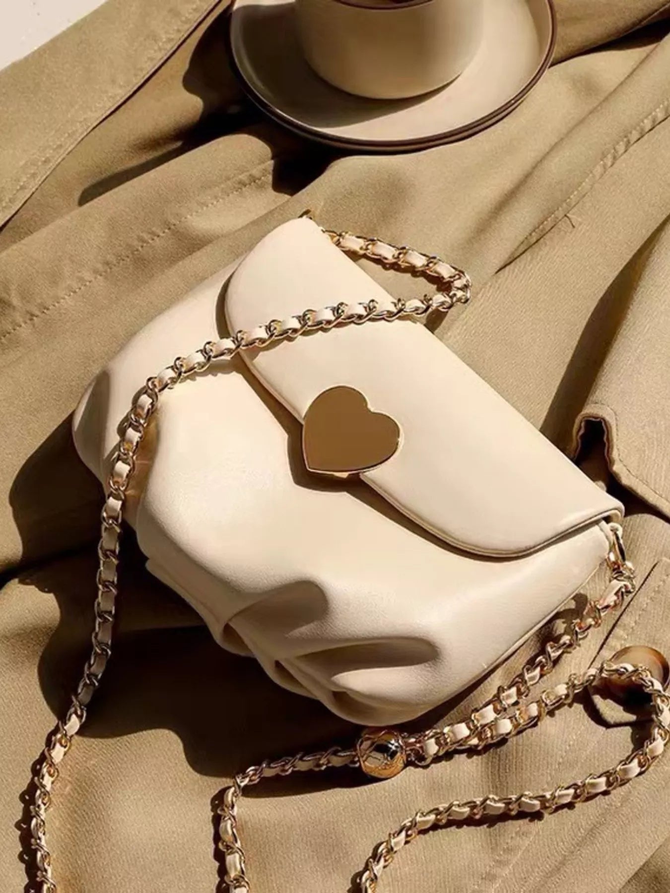 Cream mini crossbody bag with gold heart clasp and chain strap resting on beige fabric.