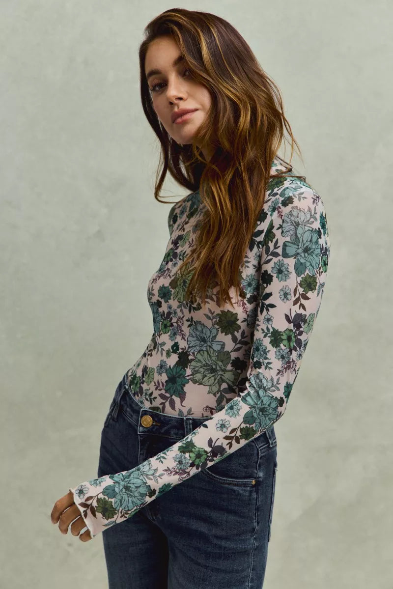 A woman wearing a sheer mesh long sleeve top with green and blue floral print tucked into dark high-waisted jeans