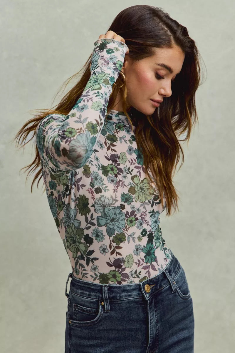 A woman wearing a sheer mesh long sleeve top with green and blue floral print tucked into dark high-waisted jeans