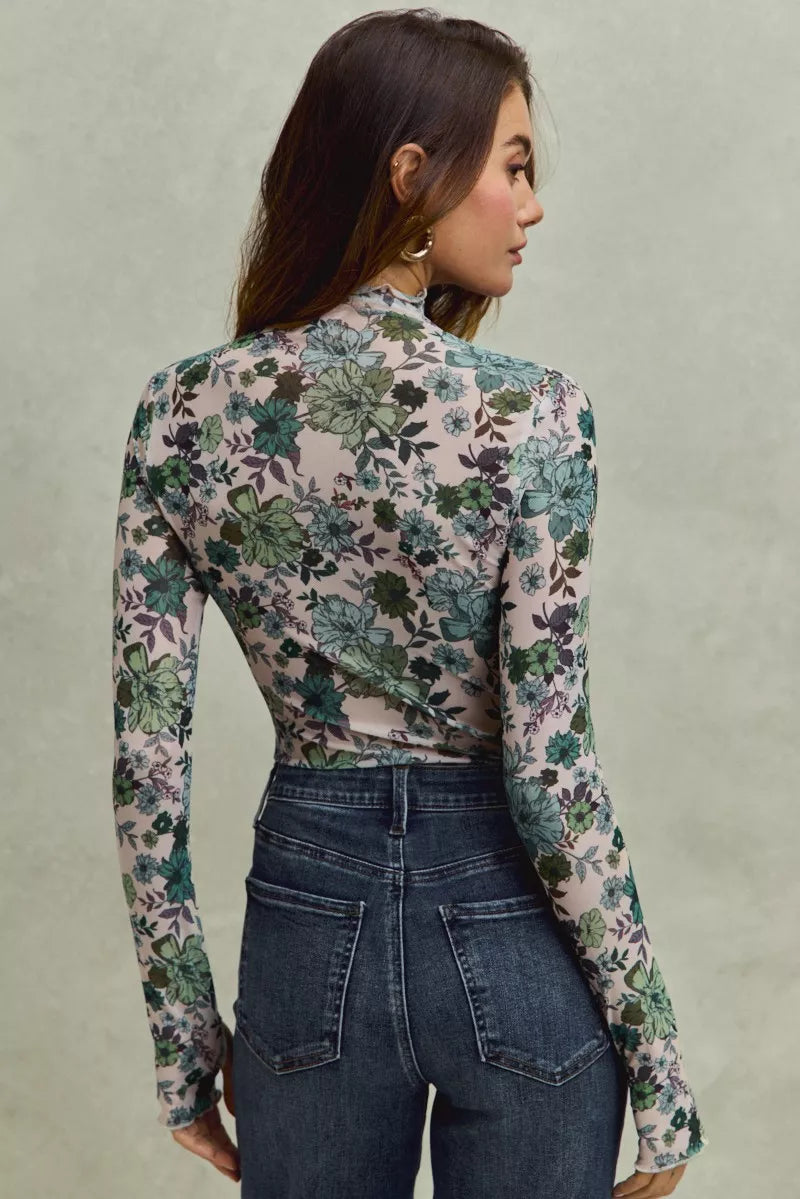 A woman wearing a sheer mesh long sleeve top with green and blue floral print tucked into dark high-waisted jeans