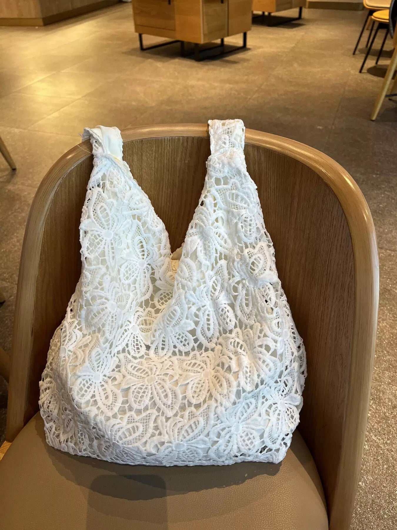 White medium-sized tote bag made of floral lace fabric with knotted straps, placed on a wooden chair in an indoor setting.