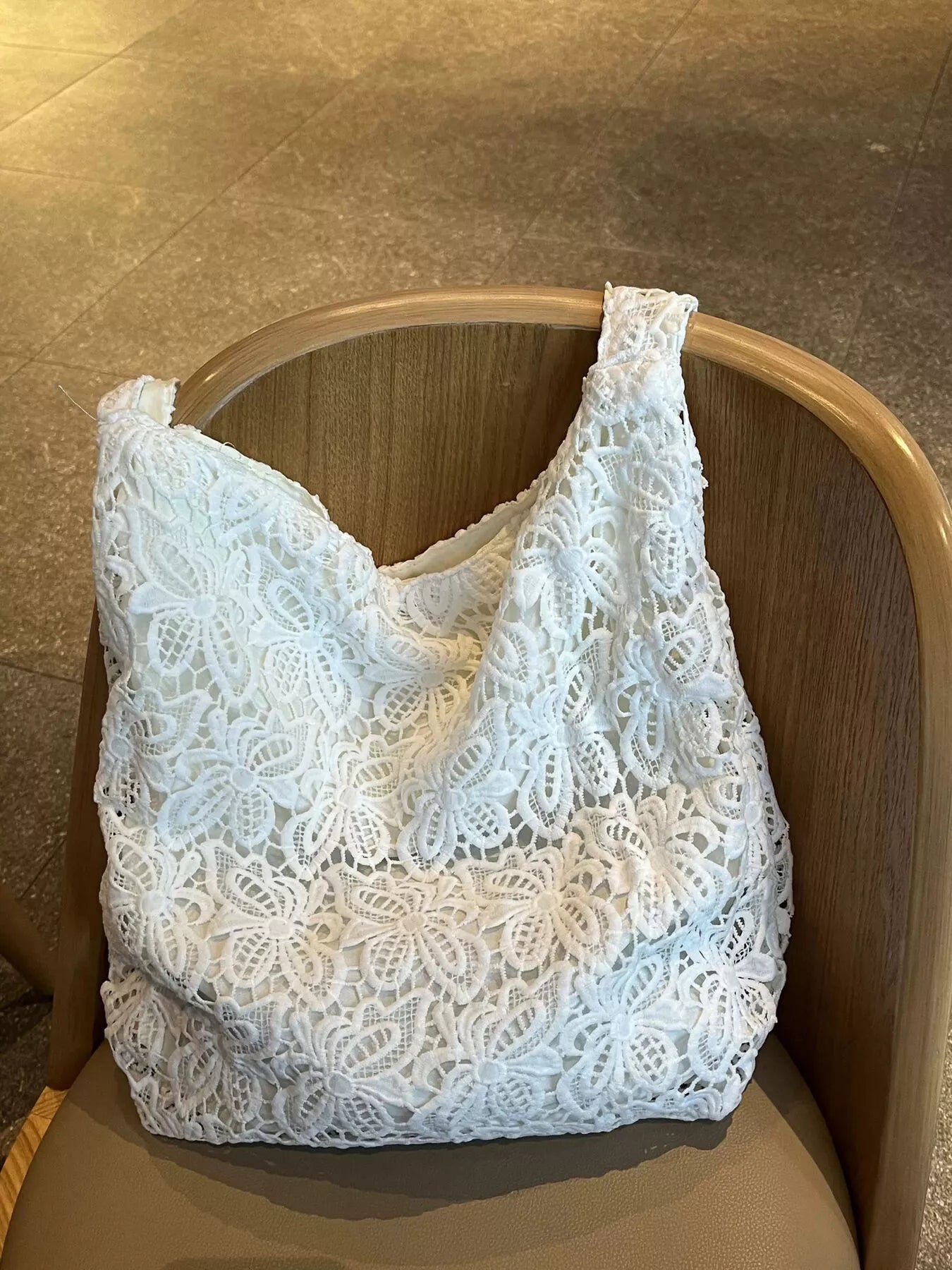 White medium-sized tote bag made of floral lace fabric with knotted straps, placed on a wooden chair in an indoor setting.