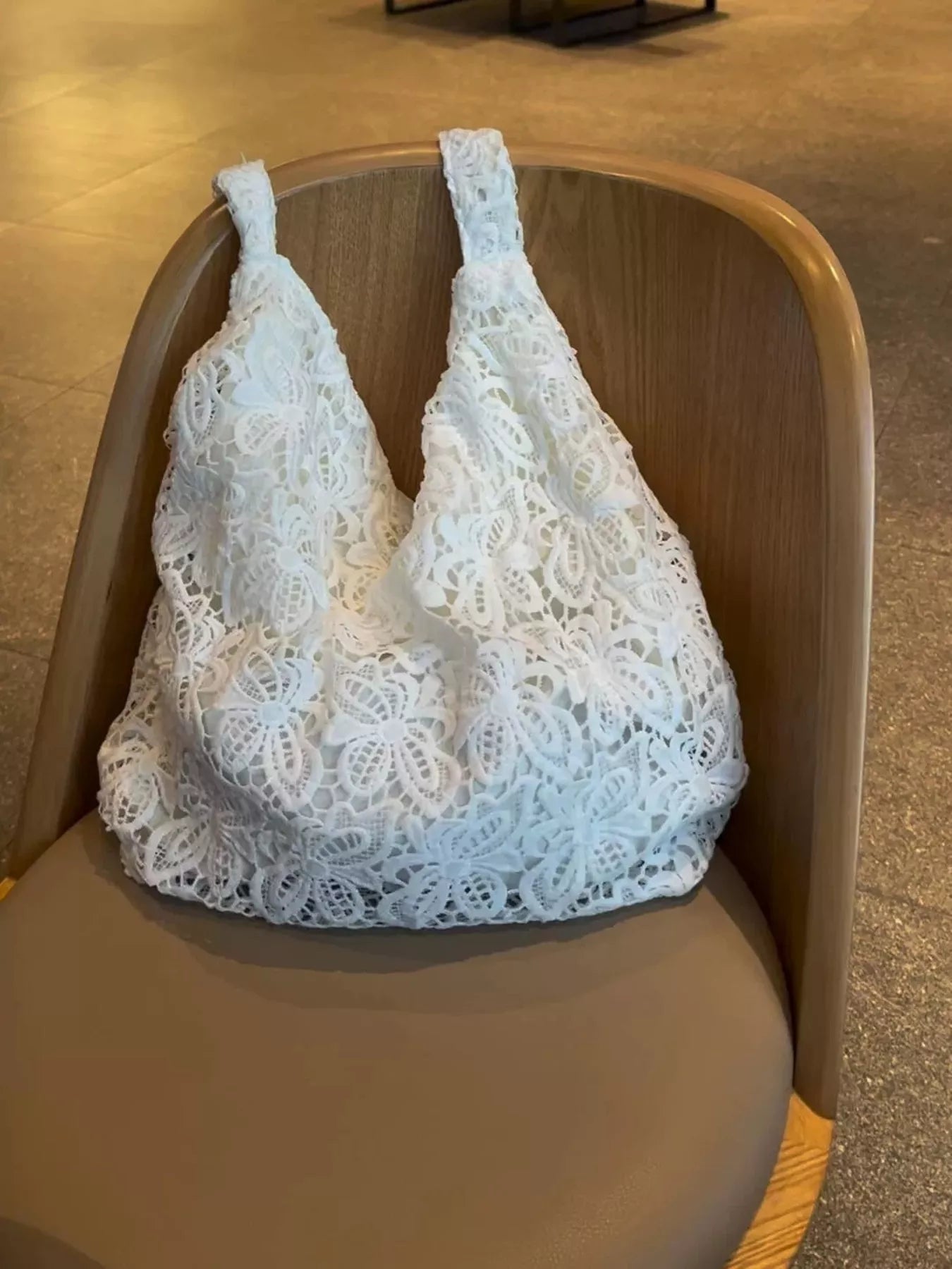 White medium-sized tote bag made of floral lace fabric with knotted straps, placed on a wooden chair in an indoor setting.