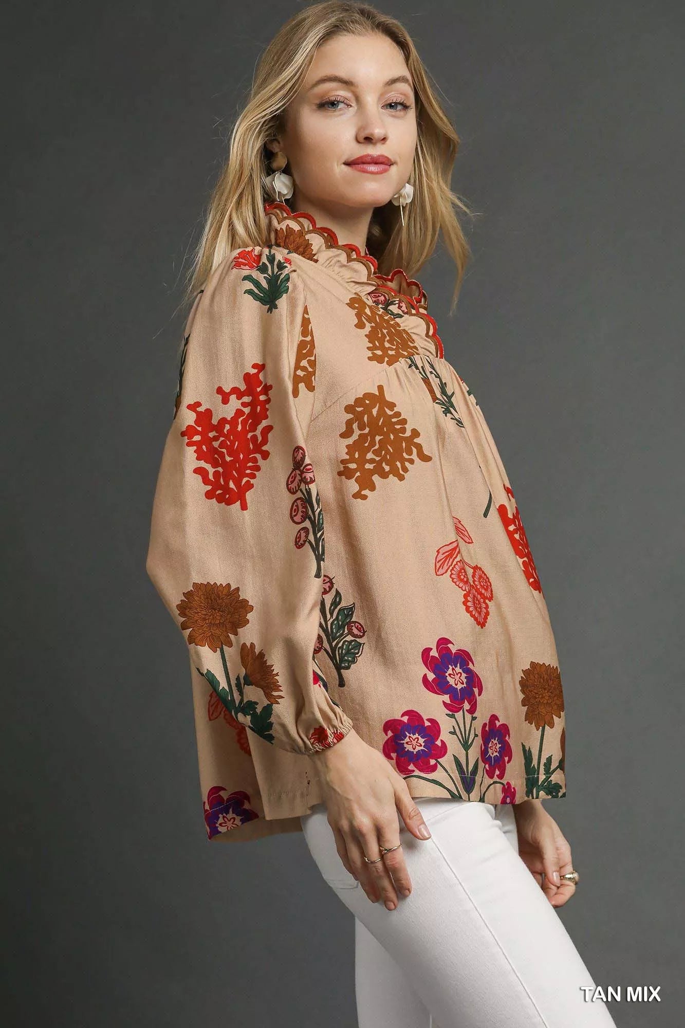 Model wearing a beige floral blouse with scalloped red-trim neckline and long puff sleeves, paired with white pants.