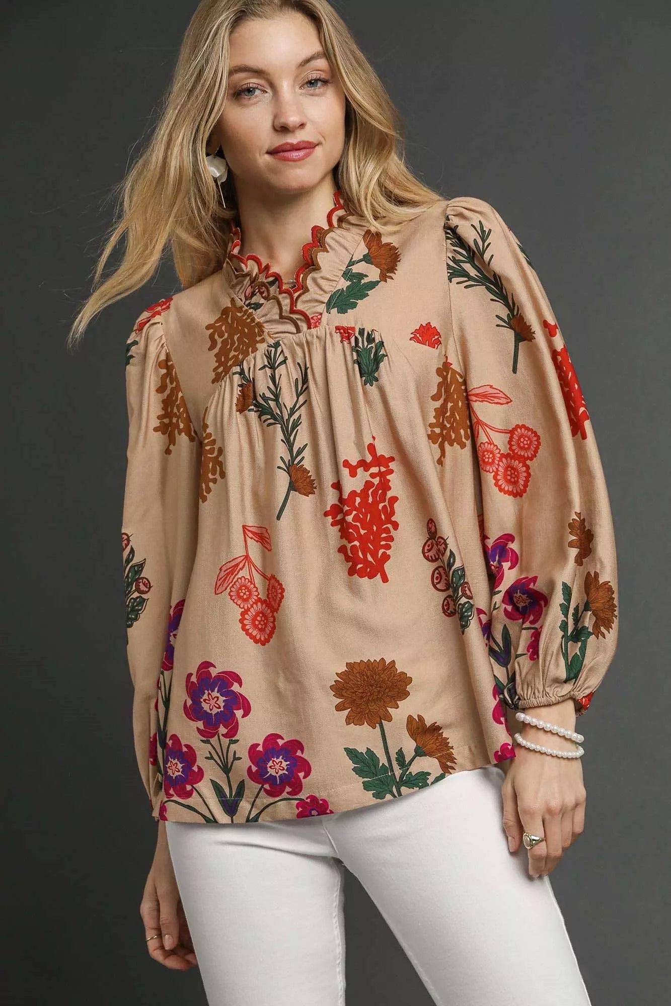 Model wearing a beige floral blouse with scalloped red-trim neckline and long puff sleeves, paired with white pants.
