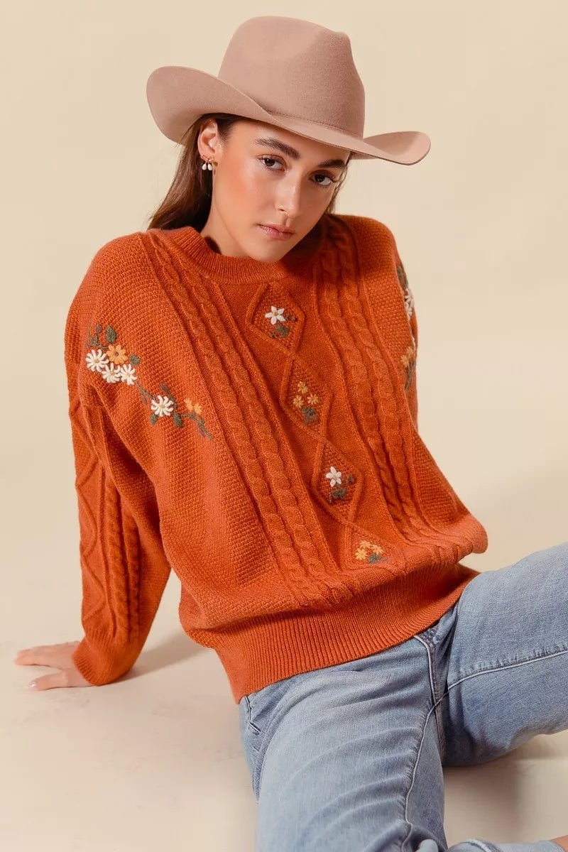 Model wearing a rust-colored cable-knit sweater with floral embroidery and relaxed fit, styled with light wash jeans and a beige hat for a cozy autumn look.