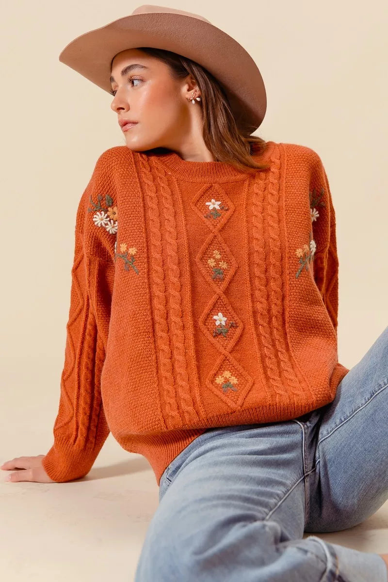 Model wearing a rust-colored cable-knit sweater with floral embroidery and relaxed fit, styled with light wash jeans and a beige hat for a cozy autumn look.