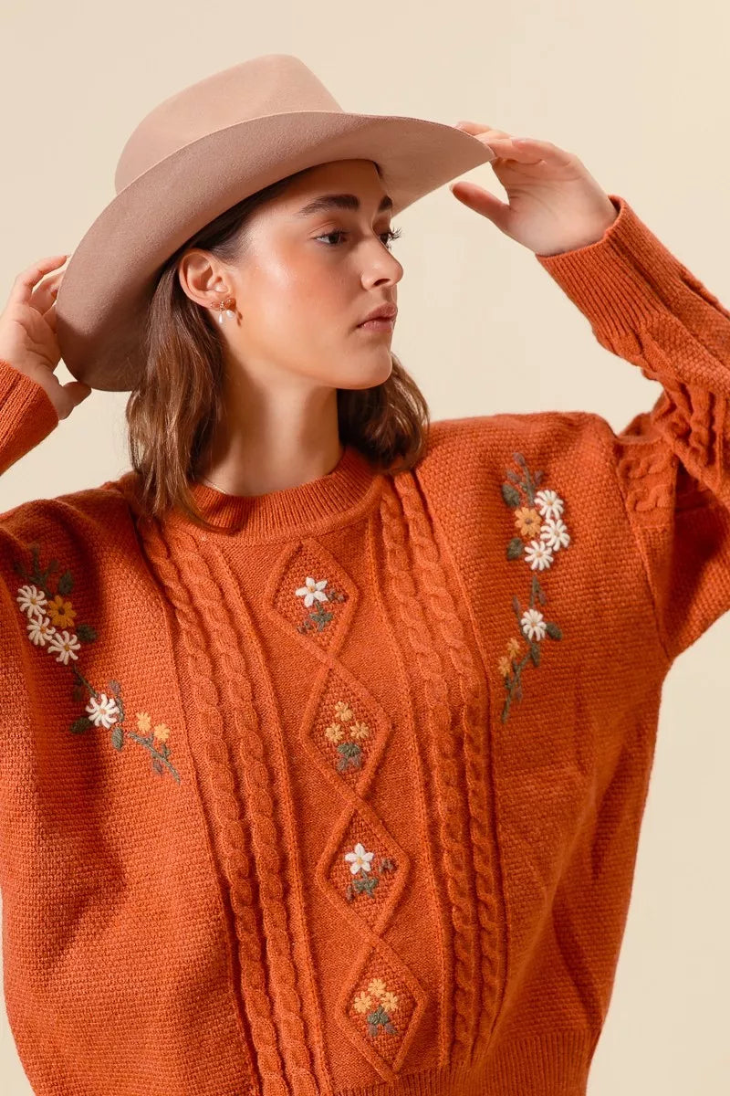 Model wearing a rust-colored cable-knit sweater with floral embroidery and relaxed fit, styled with light wash jeans and a beige hat for a cozy autumn look.