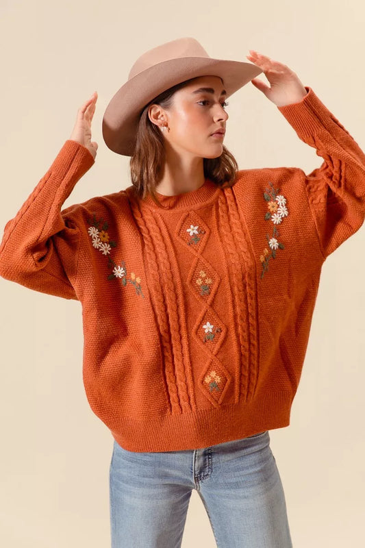 Model wearing a rust-colored cable-knit sweater with floral embroidery and relaxed fit, styled with light wash jeans and a beige hat for a cozy autumn look.