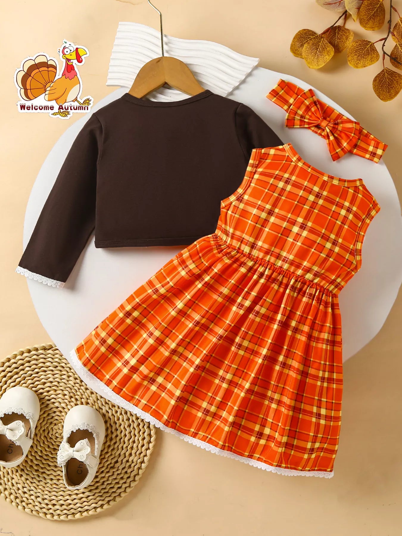 Three-piece baby girl outfit with orange plaid turkey dress, brown cardigan, and matching plaid headband on a beige background with fall leaves.