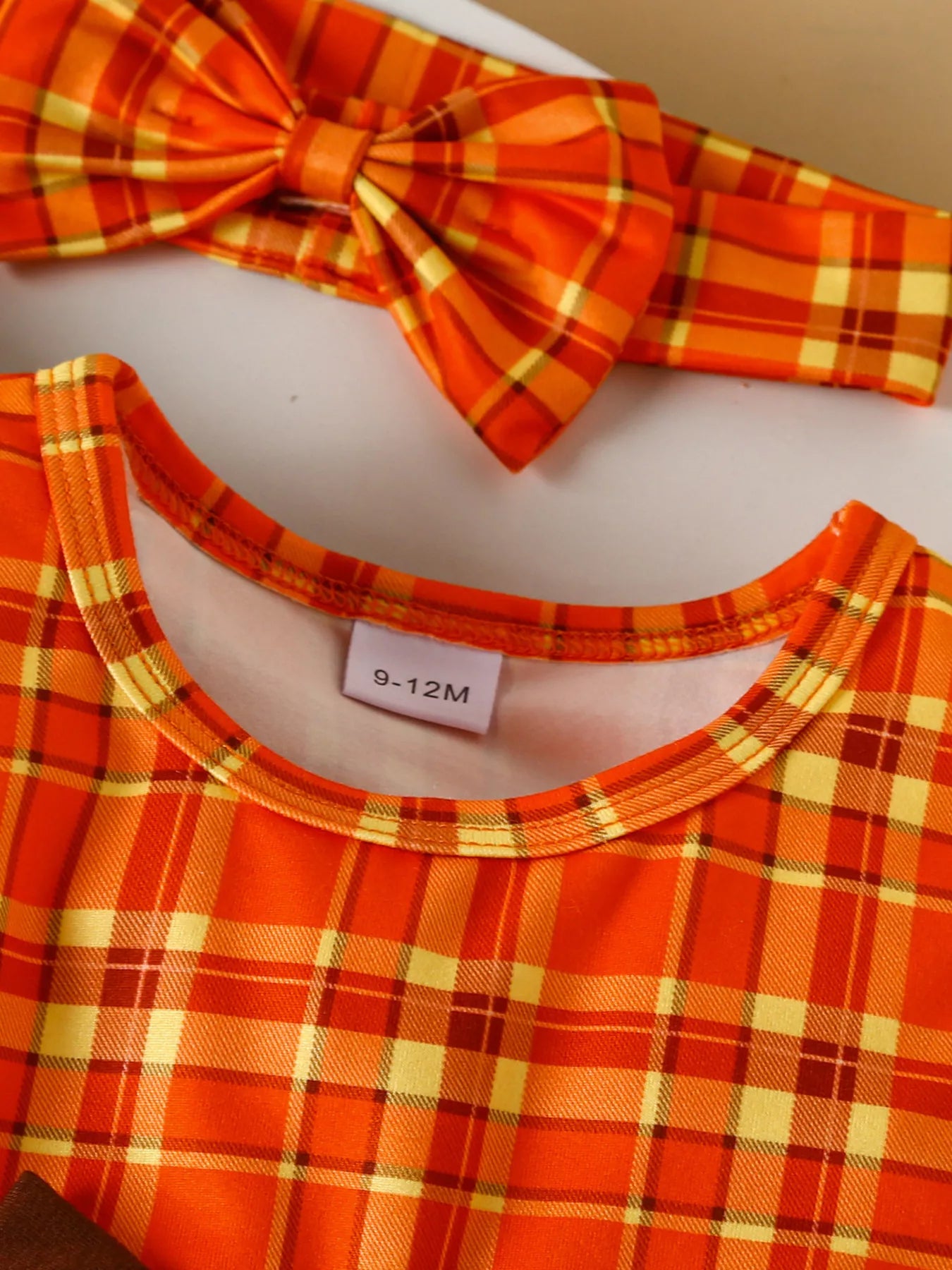 Three-piece baby girl outfit with orange plaid turkey dress, brown cardigan, and matching plaid headband on a beige background with fall leaves.