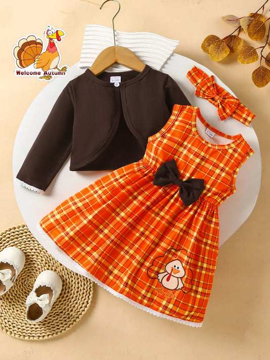 Three-piece baby girl outfit with orange plaid turkey dress, brown cardigan, and matching plaid headband on a beige background with fall leaves.