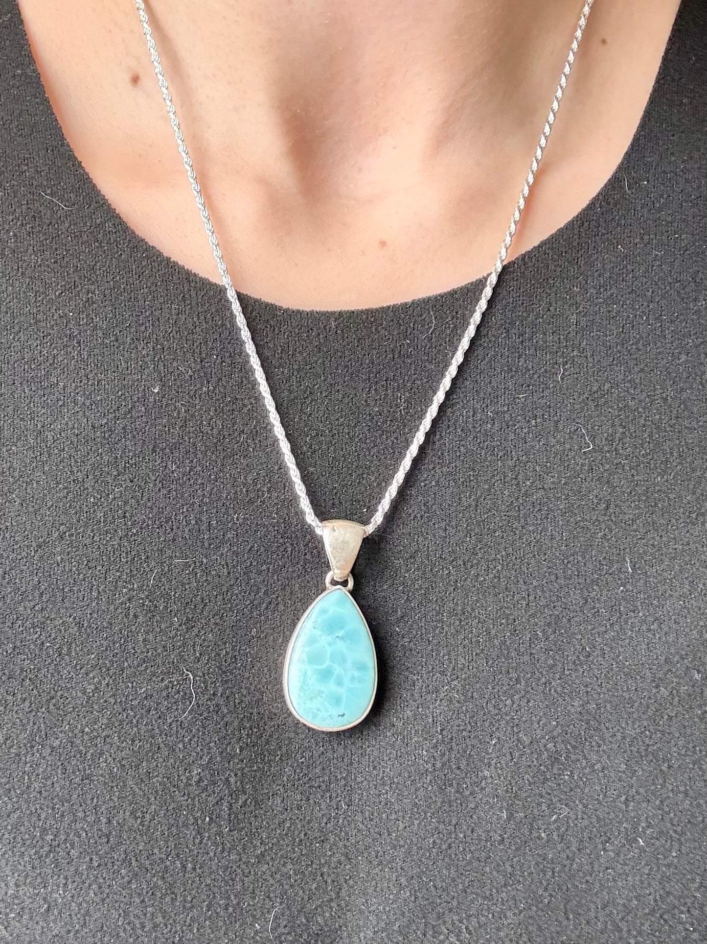 Teardrop-shaped blue larimar gemstone pendant with natural marbling on a neutral stone background