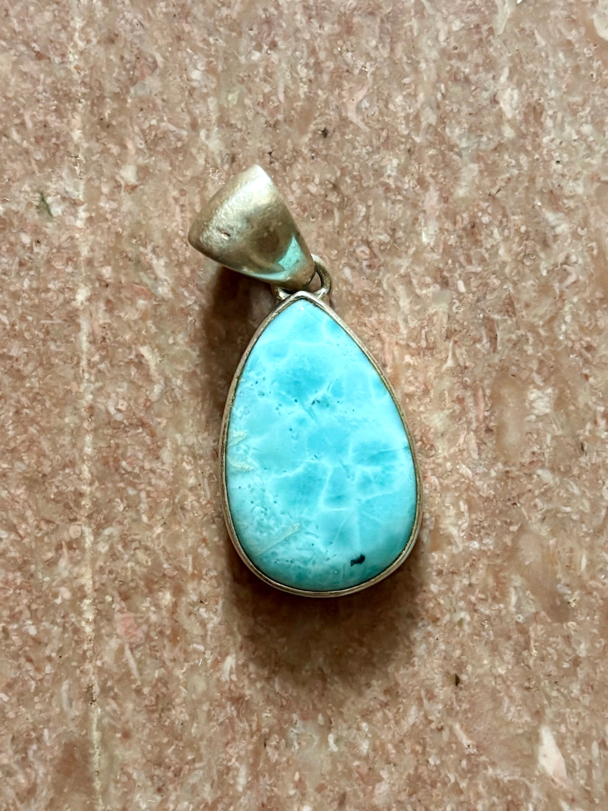 Teardrop-shaped blue larimar gemstone pendant with natural marbling on a neutral stone background