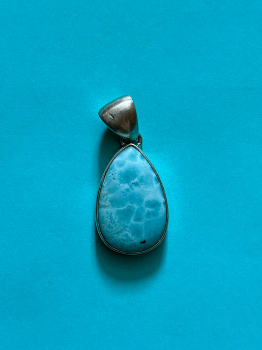 Teardrop-shaped blue larimar gemstone pendant with natural marbling on a neutral stone background