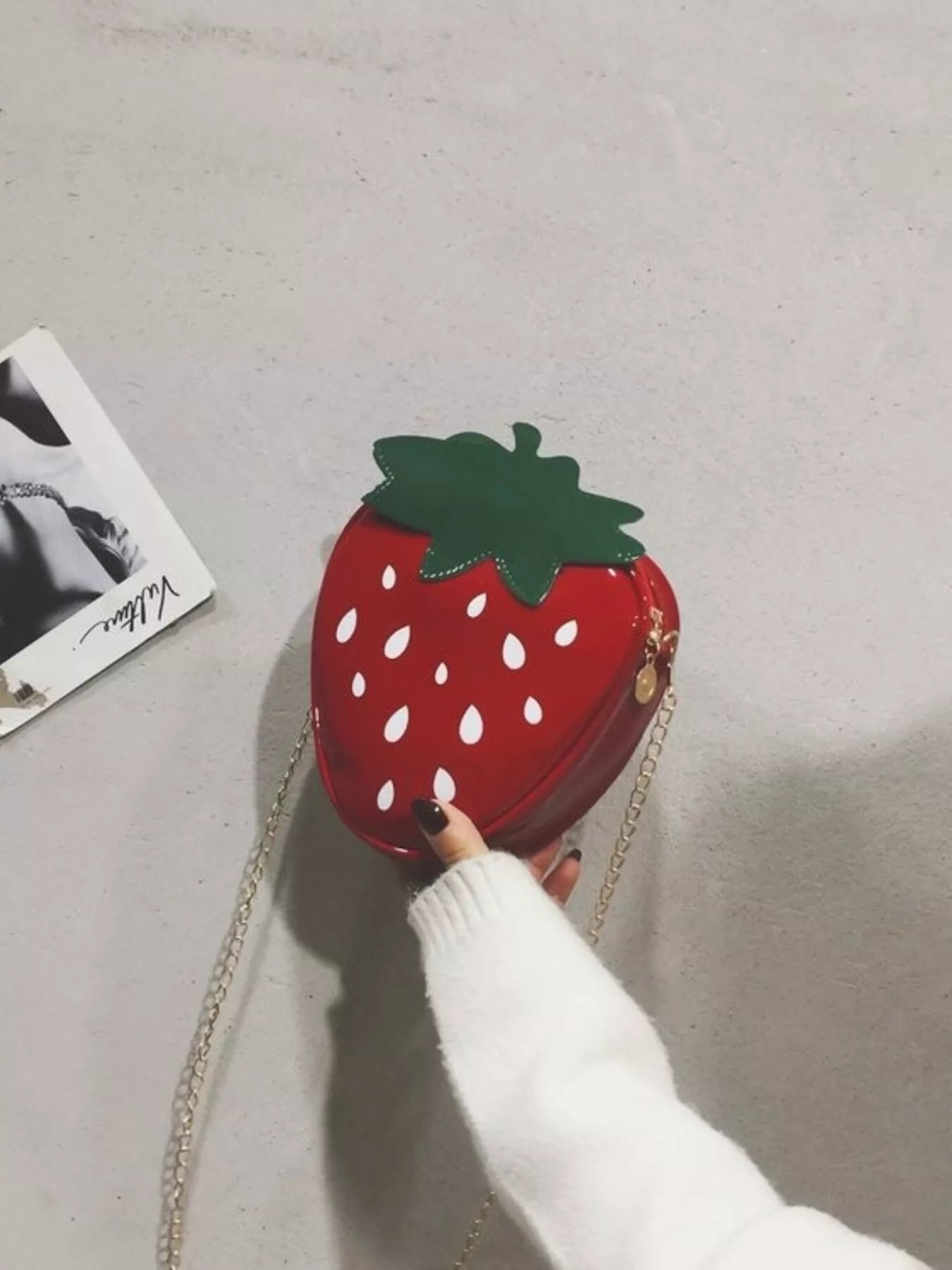 Glossy red strawberry-shaped mini bag with green leaf top and gold chain strap, displayed on a neutral background for a cute and playful look.