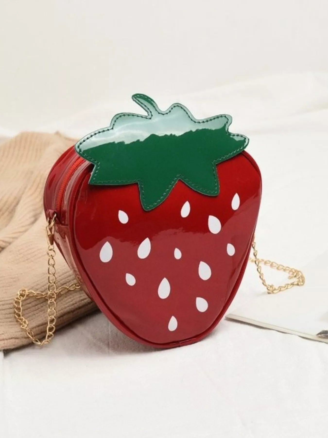 Glossy red strawberry-shaped mini bag with green leaf top and gold chain strap, displayed on a neutral background for a cute and playful look.