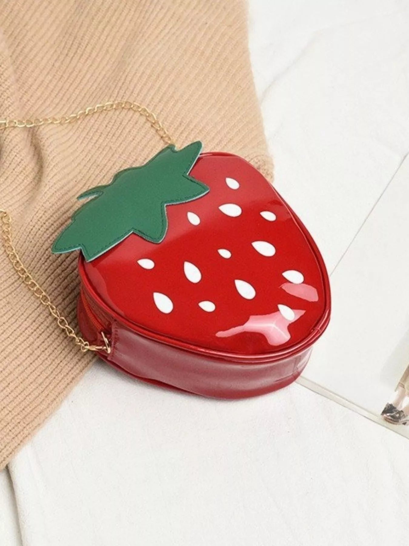Glossy red strawberry-shaped mini bag with green leaf top and gold chain strap, displayed on a neutral background for a cute and playful look.
