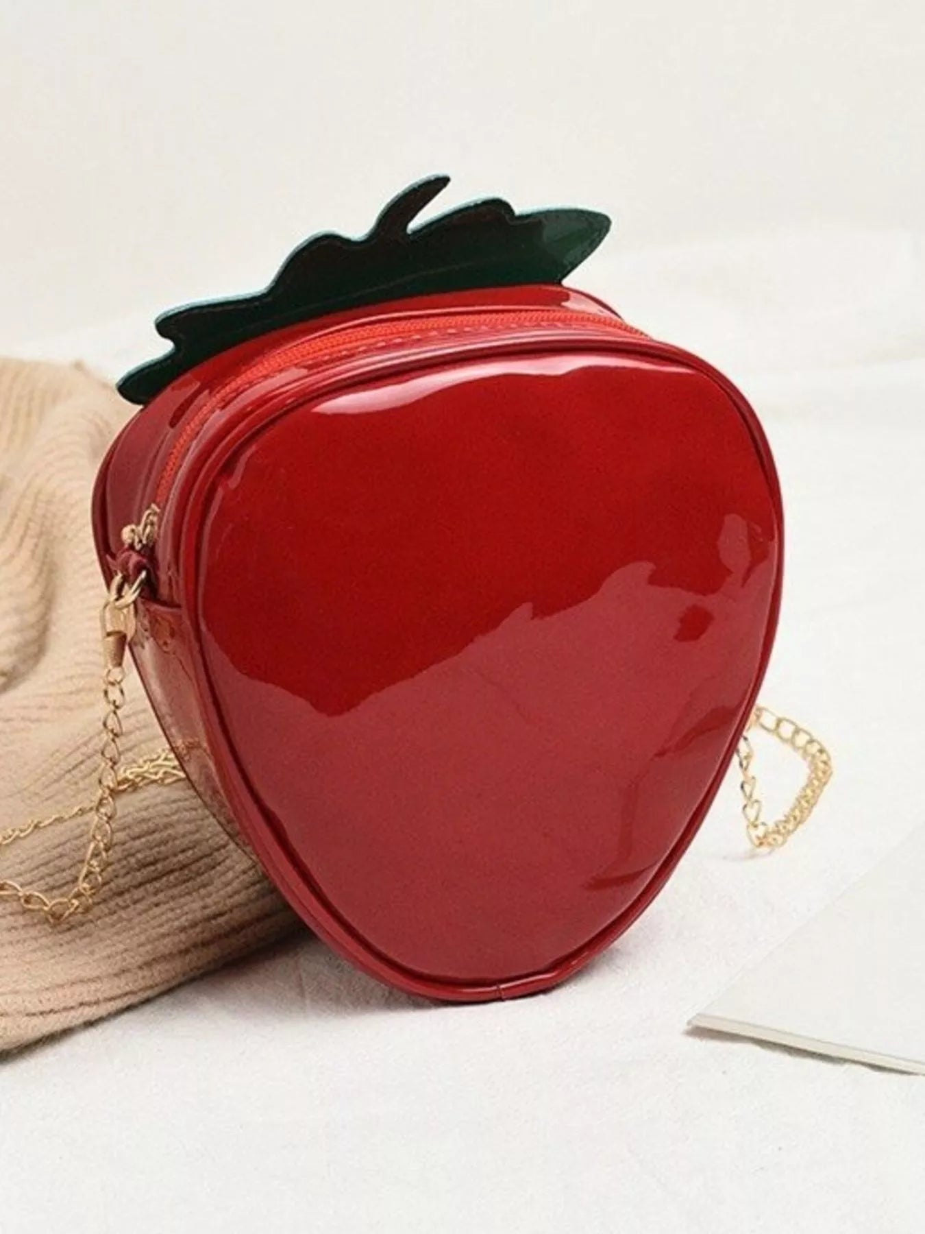 Glossy red strawberry-shaped mini bag with green leaf top and gold chain strap, displayed on a neutral background for a cute and playful look.