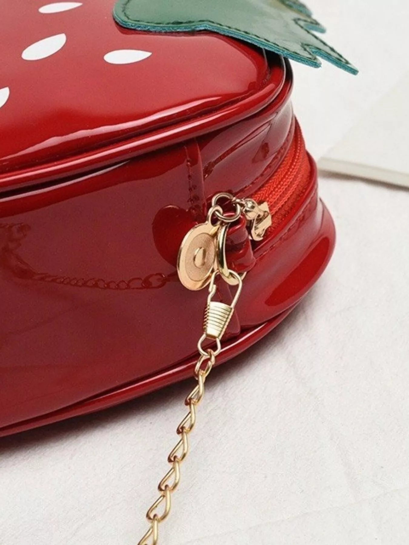 Glossy red strawberry-shaped mini bag with green leaf top and gold chain strap, displayed on a neutral background for a cute and playful look.