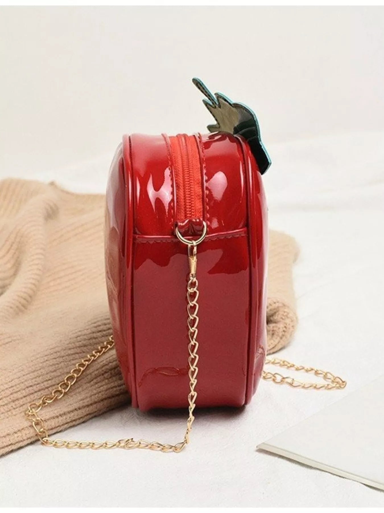 Glossy red strawberry-shaped mini bag with green leaf top and gold chain strap, displayed on a neutral background for a cute and playful look.