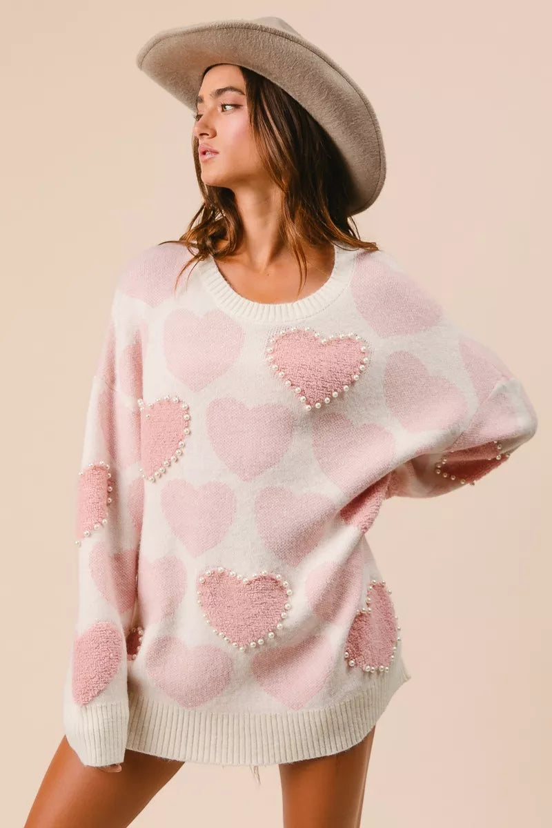 Woman wearing a cream knit sweater with pink heart pattern and pearl embellishments
