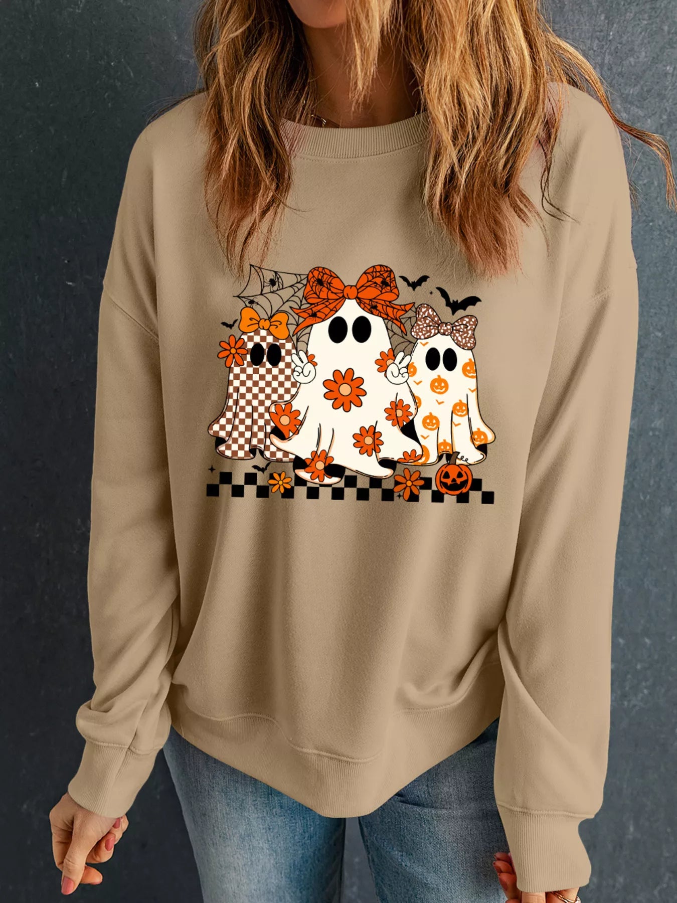 Woman wearing a tan crewneck sweatshirt with three cute boho-style ghosts featuring bows and floral pumpkin patterns — Halloween sweatshirt from A Whimsical Affair.