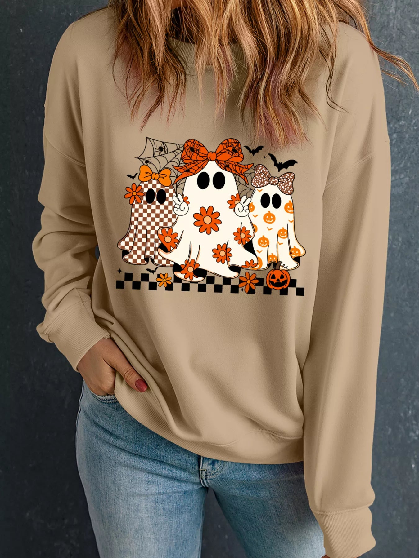 Woman wearing a tan crewneck sweatshirt with three cute boho-style ghosts featuring bows and floral pumpkin patterns — Halloween sweatshirt from A Whimsical Affair.