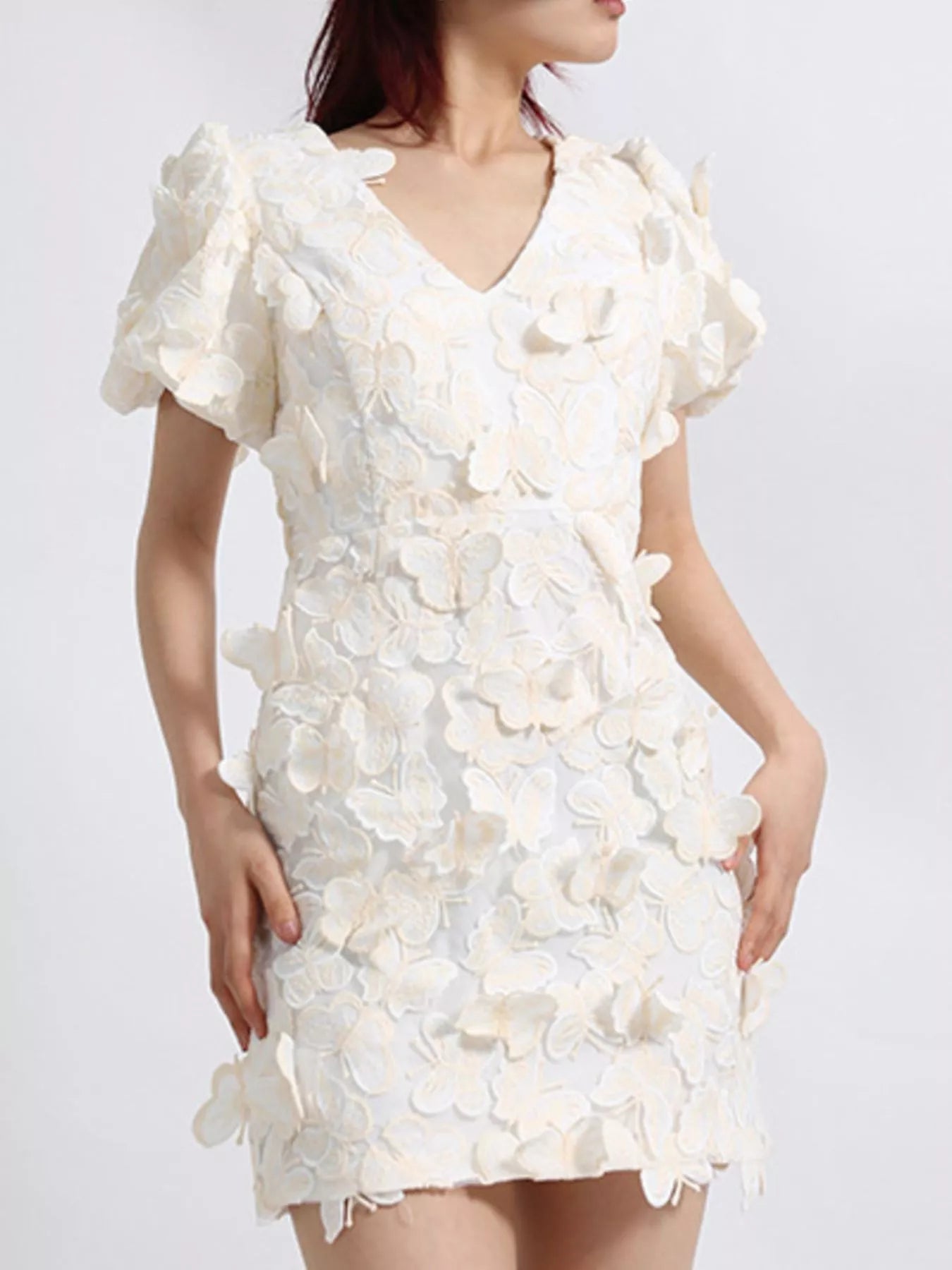 Model wearing a white mini dress adorned with 3D butterfly appliqués, featuring short puff sleeves, a V-neckline, and a fitted silhouette, styled against a plain background.