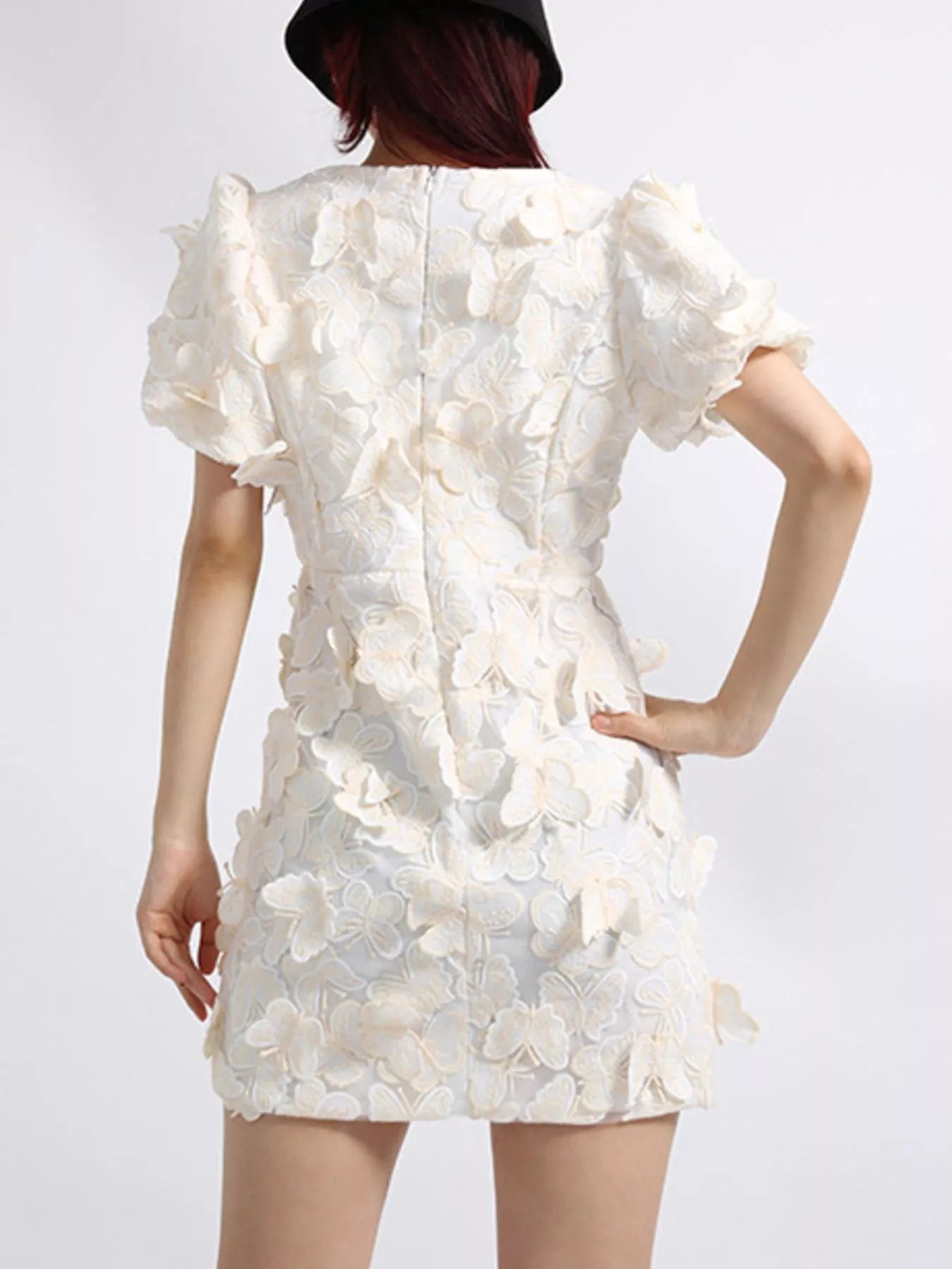 Model wearing a white mini dress adorned with 3D butterfly appliqués, featuring short puff sleeves, a V-neckline, and a fitted silhouette, styled against a plain background.