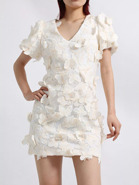 Model wearing a white mini dress adorned with 3D butterfly appliqués, featuring short puff sleeves, a V-neckline, and a fitted silhouette, styled against a plain background.