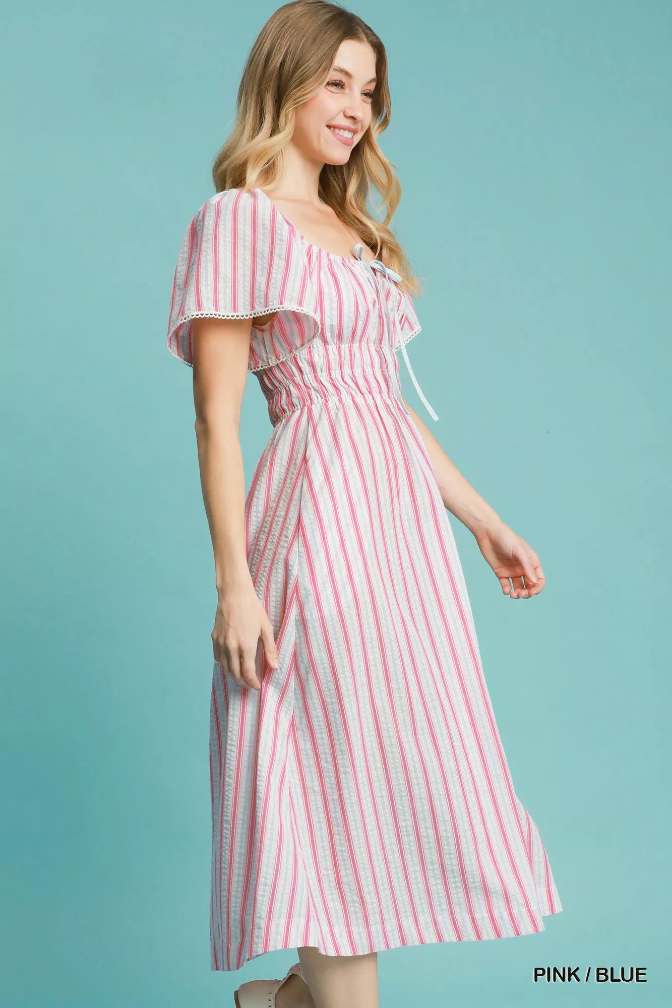Model wearing a pink-and-blue striped smocked midi dress with ruffled short sleeves, paired with neutral flats and a pink crossbody bag.