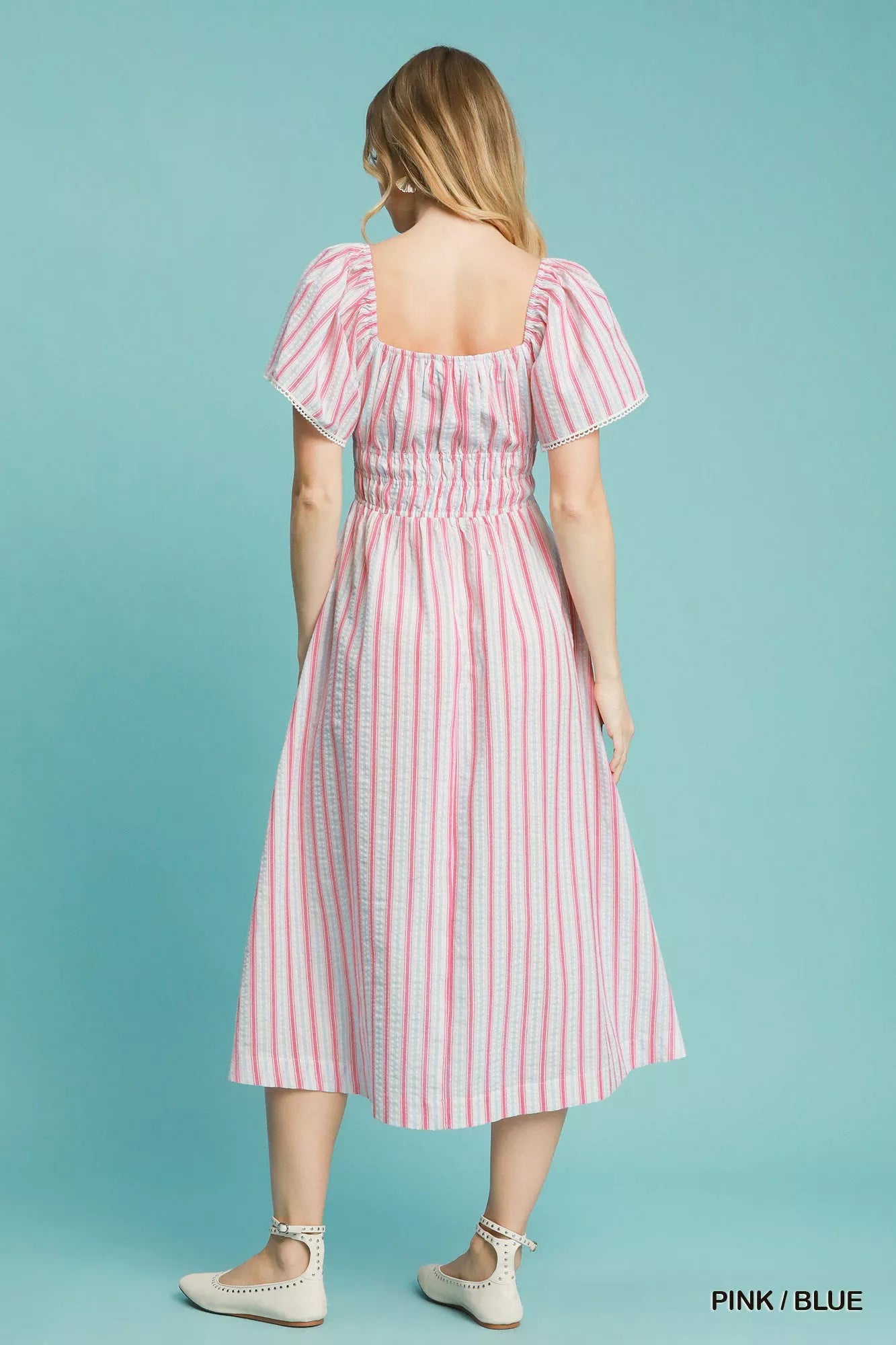 Model wearing a pink-and-blue striped smocked midi dress with ruffled short sleeves, paired with neutral flats and a pink crossbody bag.