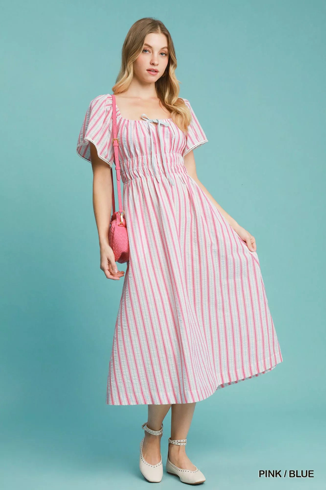 Model wearing a pink-and-blue striped smocked midi dress with ruffled short sleeves, paired with neutral flats and a pink crossbody bag.