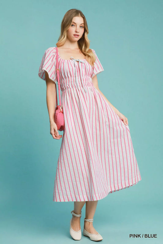 Model wearing a pink-and-blue striped smocked midi dress with ruffled short sleeves, paired with neutral flats and a pink crossbody bag.