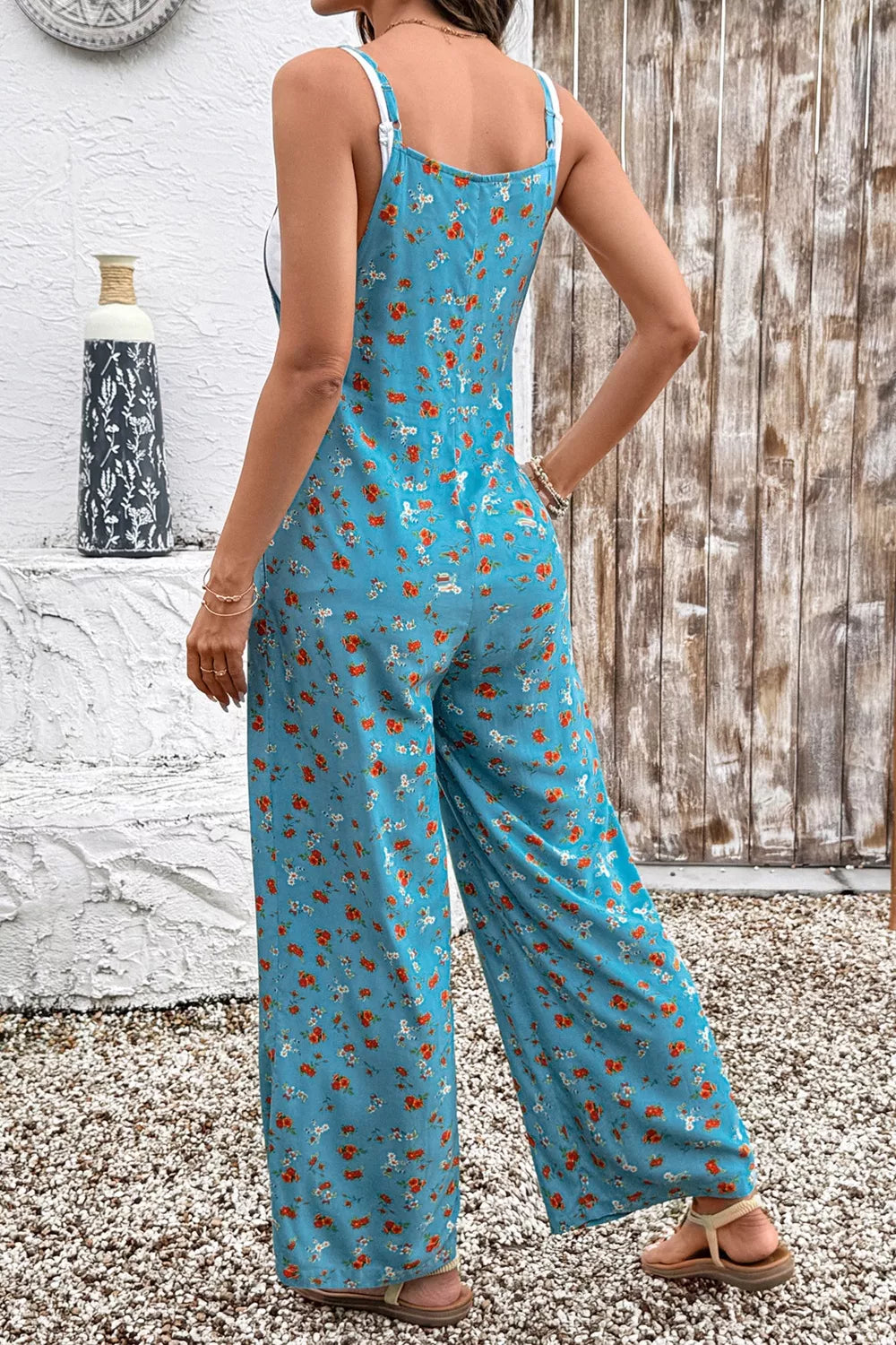 Blue floral wide-leg jumpsuit with thin straps, relaxed flowy fit, and side pockets styled with sandals.