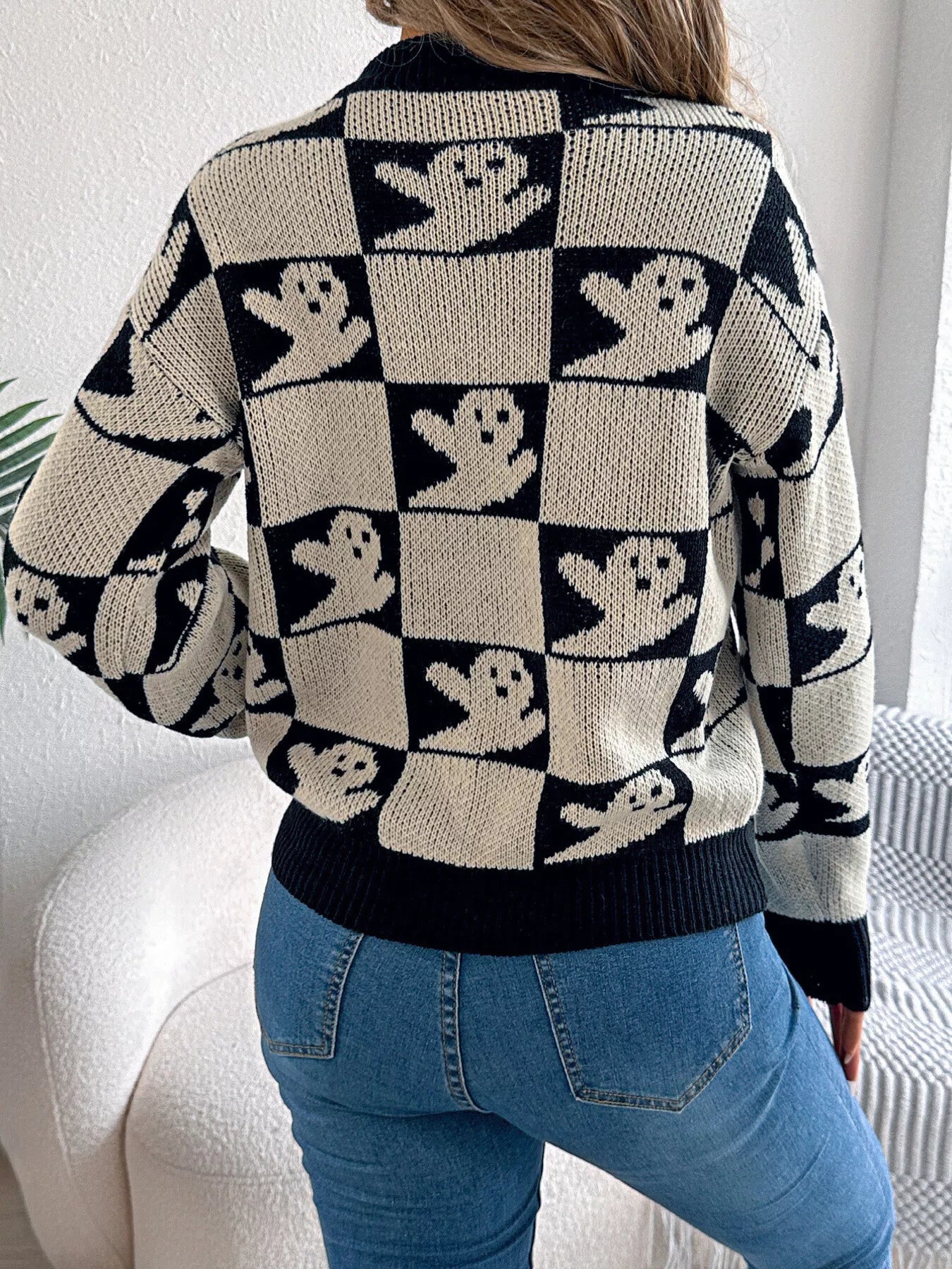 Woman wearing a black checkerboard knit sweater with ghost pattern, styled with jeans and a white purse — cozy Halloween sweater from A Whimsical Affair.