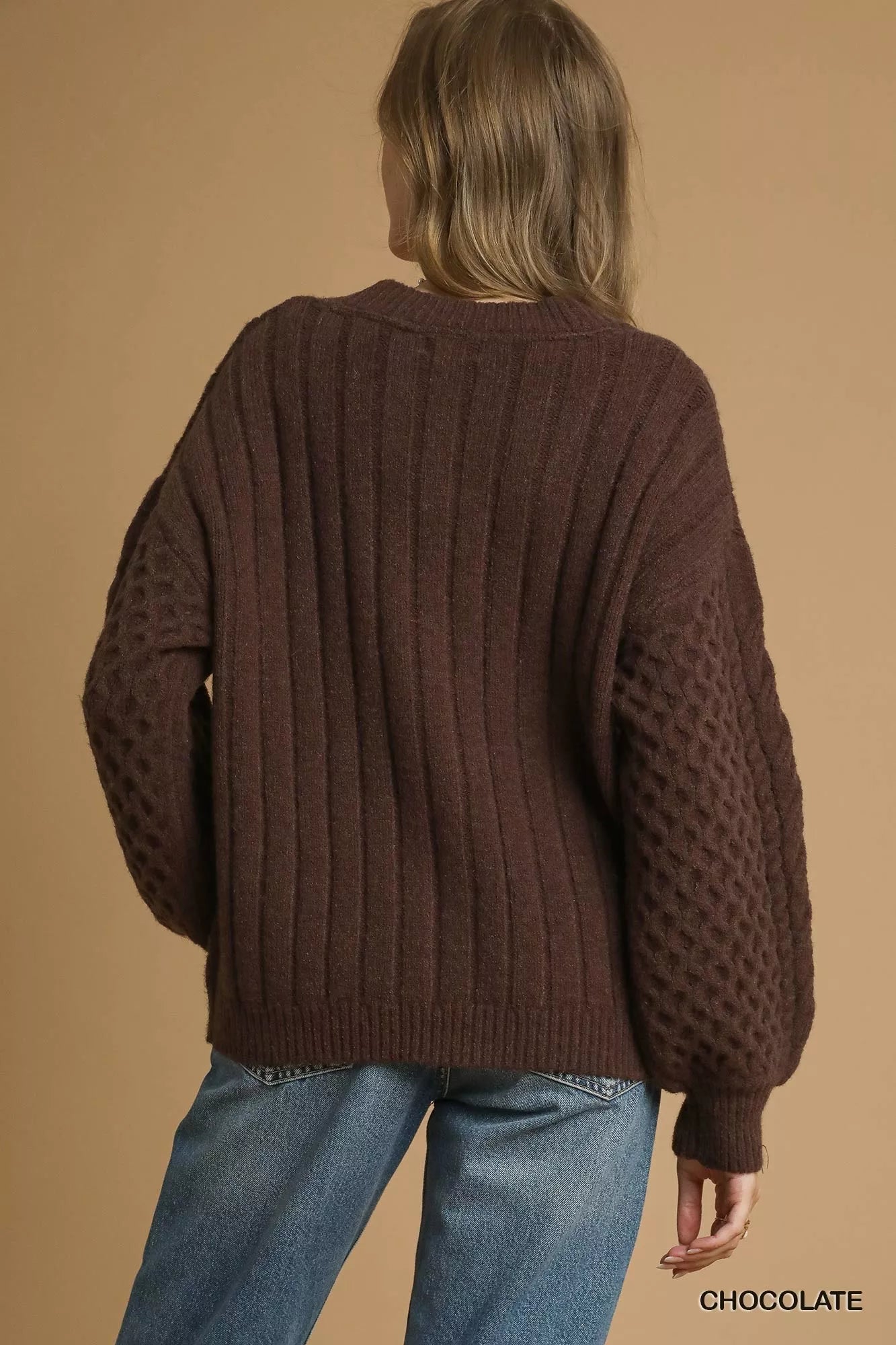Model wearing a chocolate-brown cable-knit cardigan with pearl buttons, styled with a white tank top and jeans.